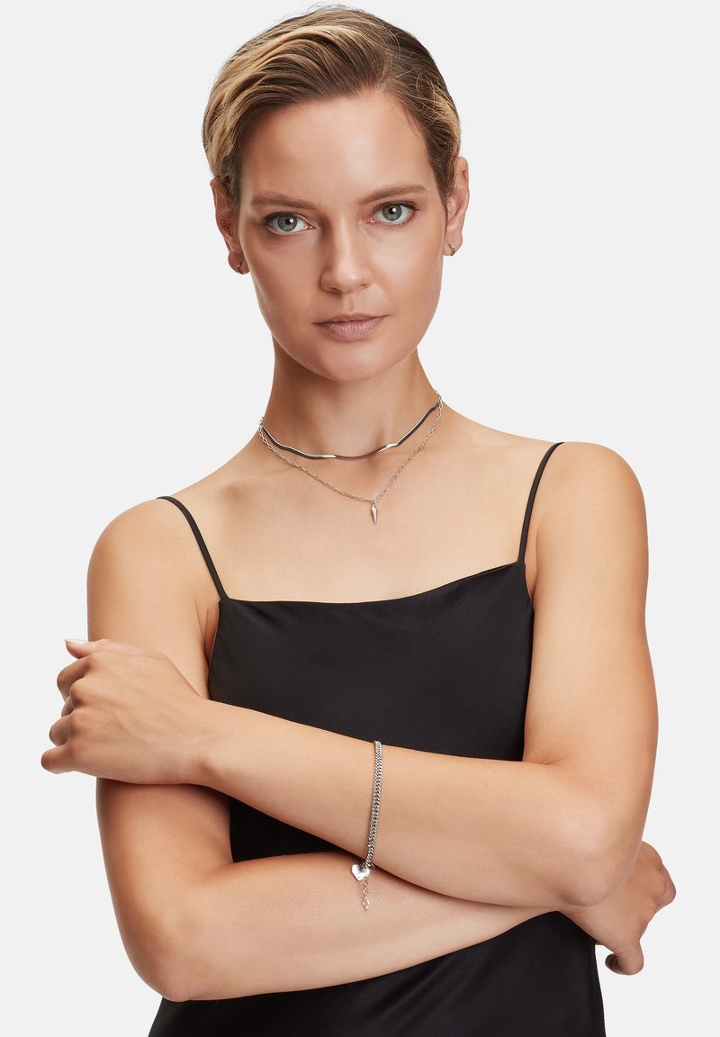 Silberne Halsketten, die schichtweise um den Hals getragen werden, mit einem wellenförmigen Design und einem spitzen Anhänger; ein silbernes Armband mit einem Herzanhänger am Handgelenk.