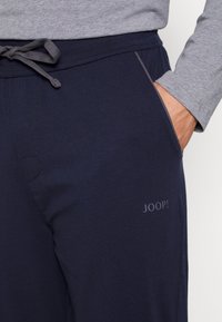 Marineblå joggebukser laget av myk stoff med elastisk midje og snor. Har en sidelomme og en subtil "JOOP!" logo.