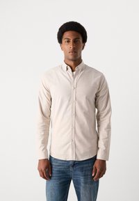 Μπεζ πουκάμισο με κουμπιά φτιαγμένο από βαμβάκι, με στενό γιακά, μακριά μανίκια και λεία υφή, σε συνδυασμό με μπλε τζιν.