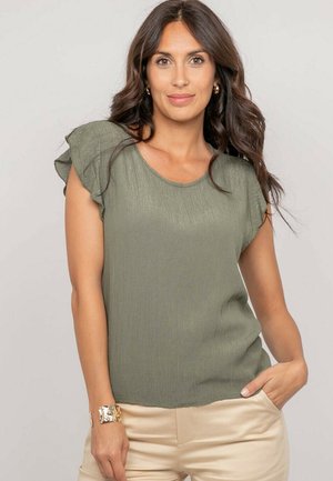 Femme aux longs cheveux noirs portant des boucles d'oreilles anneaux dorées, un haut à manches courtes vert olive, un pantalon beige et un bracelet doré, debout avec une main dans la poche.