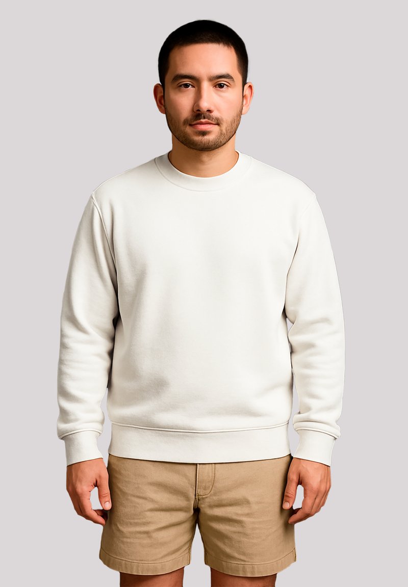 Man with short dark hair wearing a plain white sweatshirt and tan shorts standing against a neutral light gray background.