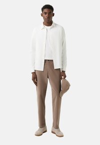 Hvid skjorte over en hvid T-shirt, brune bukser og beige sneakers. Holder en lysebrun kasket. Enkel, ren design.