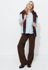 Chemise bleu clair à boutons, gilet en tricot marron drapé sur une épaule, pantalon pattes d'éléphant marron scintillant et bottes à plateforme noires.