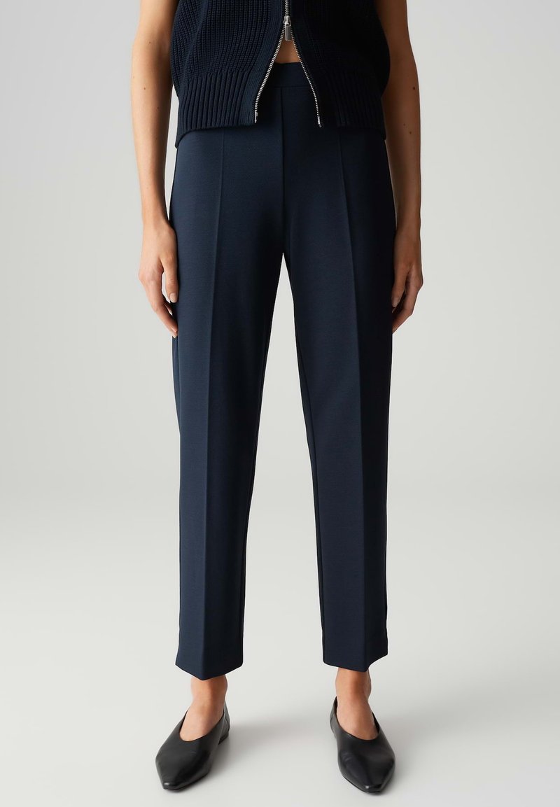 Pantalons navy ajustés en tissu lisse, présentant un design à jambes droites avec des plis à l'avant et des chevilles ourlées, associés à des ballerines noires.