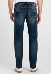 Jeans de mezclilla azul oscuro con detalles de desgaste, corte slim, bolsillos traseros con solapas en ángulo y un parche de marca de cuero marrón en la cintura.