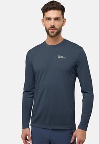 Un homme aux cheveux bruns courts et à la barbe, portant une chemise à manches longues bleu marine avec le logo Jack Wolfskin sur la poitrine, se tenant devant un fond blanc.