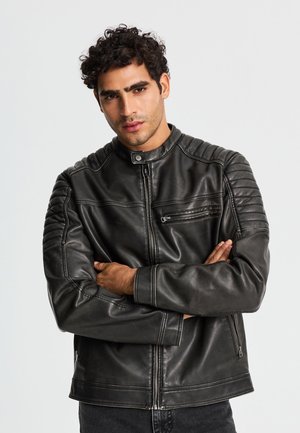 Uomo con capelli ricci che indossa una giacca di pelle nera con dettagli trapuntati sulle spalle, in piedi con le braccia incrociate davanti a uno sfondo semplice.