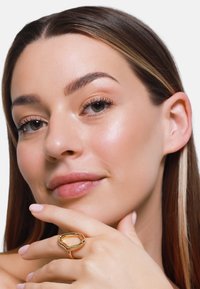 Bague en métal doré avec une forme organique sur une main, présentant une finition brillante, une texture lisse et de nombreux détails des doigts sur un fond neutre.