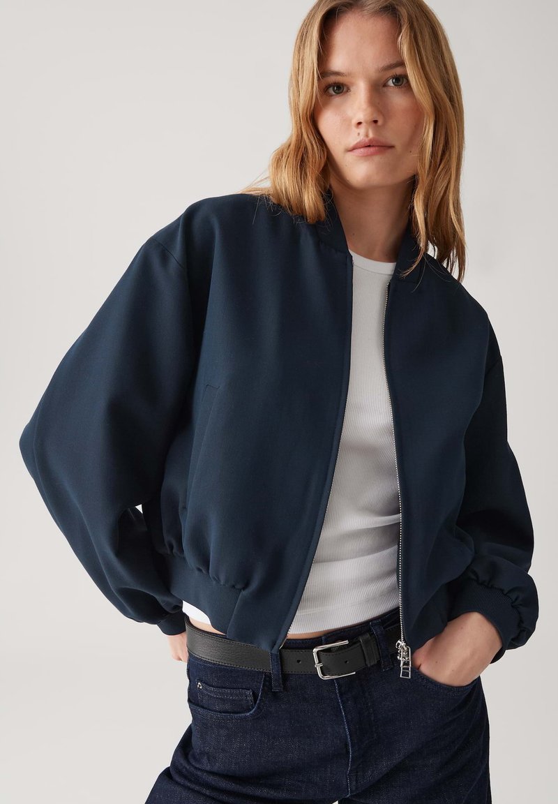 Giacca bomber blu navy con polsini e orlo elasticizzati, dotata di una cerniera frontale e tessuto liscio, abbinata a una maglietta bianca e jeans scuri.