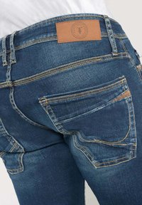 Jeans in denim di colore blu medio, design a cinque tasche, cuciture gialle a contrasto e un'etichetta in pelle marrone sulla vita.