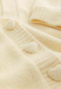 Krēmkrāsas adīts džemperis ar mīkstu tekstūru, kas rotāts ar sirds formas detaļām un ribotiem malām. Omulīgas un pievilcīgas dizaina iezīmes.