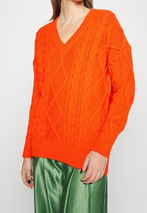 Woman wearing a bright orange cable-knit V-neck sweater and shiny green satin pants, standing with arms relaxed by sides.