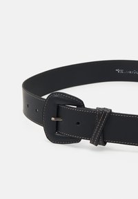 Curea neagră din piele cu cataramă rotunjită, cusături bej de-a lungul marginilor și o buclă care fixează capătul curelei pe un fundal simplu.