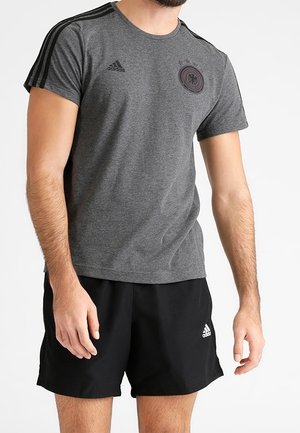 Graues Sport-T-Shirt mit schwarzen Streifen an den Schultern, Markenlogo und Emblem. Kombiniert mit schwarzen Shorts, die ein weißes Logo haben.