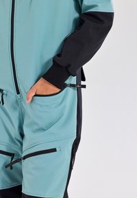 A teal and black jumpsuit features a smooth texture, zippered pockets, and elastic cuffs. The design combines durability with a modern look.
