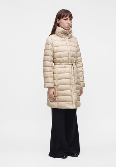 Γυναίκα που στέκεται φορώντας ένα μπεζ καπιτονέ μπουφάν με ζώνη, μαύρο φαρδύ παντελόνι και μαύρα παπούτσια σε απλό ανοιχτό φόντο.