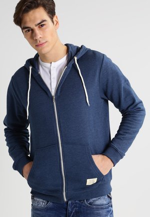 Sudadera con cremallera de color azul marino con cordones blancos, bolsillos frontales y una tela texturizada. Presenta una pequeña etiqueta de la marca en el dobladillo.