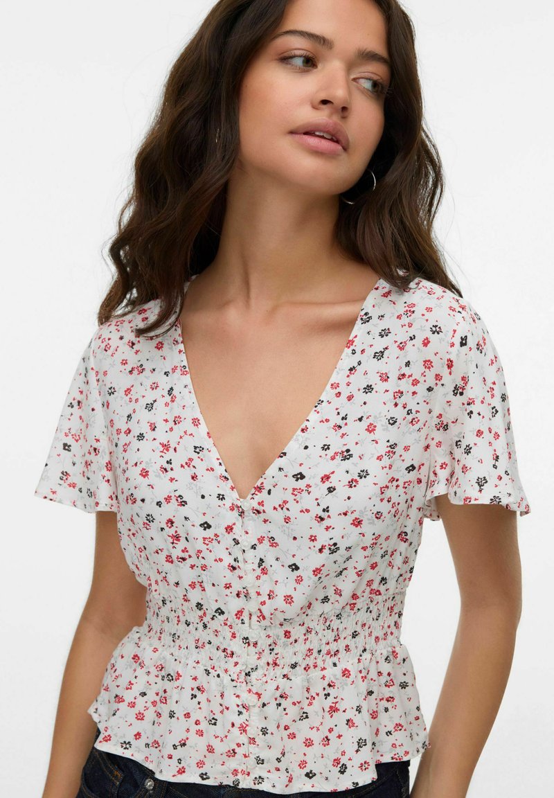 Ung kvinna som bär en vit kortärmad blus med röd och svart blommönster och knapprad, ser åt sidan.