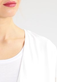 Bílá blůza s jemnou, splývavou tkaninou. Kulatý výstřih a mírně volný střih, který zdůrazňuje hladkou texturu a čisté linie.