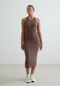 Brown ribbed tank dress with a fitted silhouette. Model wears white sneakers and pink sunglasses. Smooth texture, knee-length design.