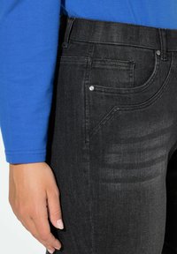 Jeans de mezclilla negra con diseño ajustado, sutil desgaste y un detalle de un solo remache metálico cerca del bolsillo delantero.