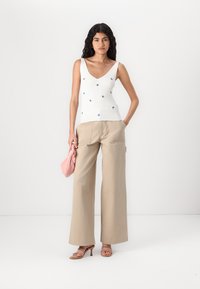 Witte geribde tanktop met kleine blauwe bloemenborduring, gecombineerd met beige wijde broek en een roze handtas. Beige sleehakken maken de look compleet.