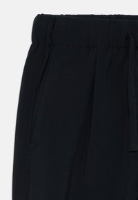 Pantalons noirs à taille élastique avec des poches latérales et un cordon de serrage sur le côté droit, présentés sur un fond blanc.
