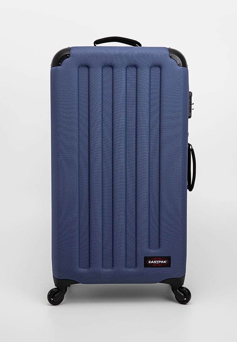 Valise rigide bleue avec des nervures verticales, des protecteurs noirs dans les coins, quatre roues pivotantes, des poignées sur le dessus et sur le côté, et un logo Eastpak.