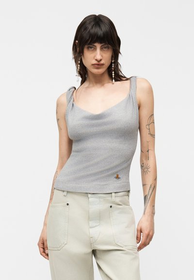 Jeune femme aux cheveux foncés et tatouages portant un haut gris sans manches et un pantalon clair, debout devant un fond blanc uni.