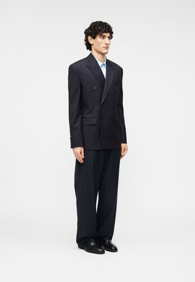 Ναυτικό μπλέ διπλόμπουτο σακάκι με πέτα με εγκοπές, συνδυασμένο με ανοιχτό μπλε πουκάμισο και παντελόνι με λεπτές ρίγες. Μαύρα δερμάτινα παπούτσια ολοκληρώνουν την εμφάνιση.