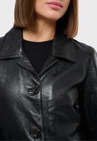 Černá kožená bunda s klasickým límcem, dvěma předními knoflíky a hladkou texturou. Přes fitted černý top.