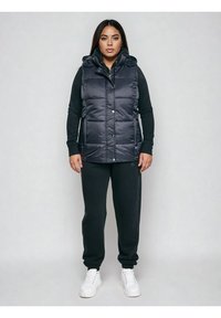 Naine seisab näoga ettepoole, kandes musta sulelist vestit, musta pika varrukaga topi, musti pükse ja valgeid tenniseid ühtlasel taustal.