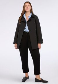 Cappotto trench nero doppiopetto con bottoni metallici, abbinato a una camicia azzurra chiara e pantaloni neri a vita alta. Mocassini neri lucidi.