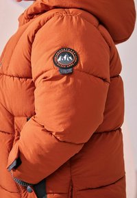 Veste orange matelassée avec un design texturé et capitonné. Elle présente un patch logo sur la manche et un accent bleu à l'ourlet et aux poches.