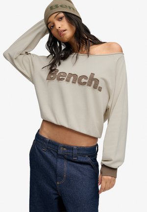 Jeune femme portant un sweat beige à épaules dénudées avec le logo "Bench." et un jean bleu, posant avec un bras levé et un bonnet beige.
