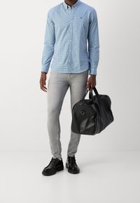 Černá kožená sportovní taška s kulatým tvarem a krátkými rukojetmi, doplněná modrou kostkovanou košilí a světle šedými úzkými džínami.