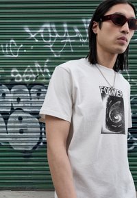 Vit bomullst-shirt med en svart spiralgrafik och ordet "FORMER." Bars med mörka solglasögon och ett silverkedjehalsband.