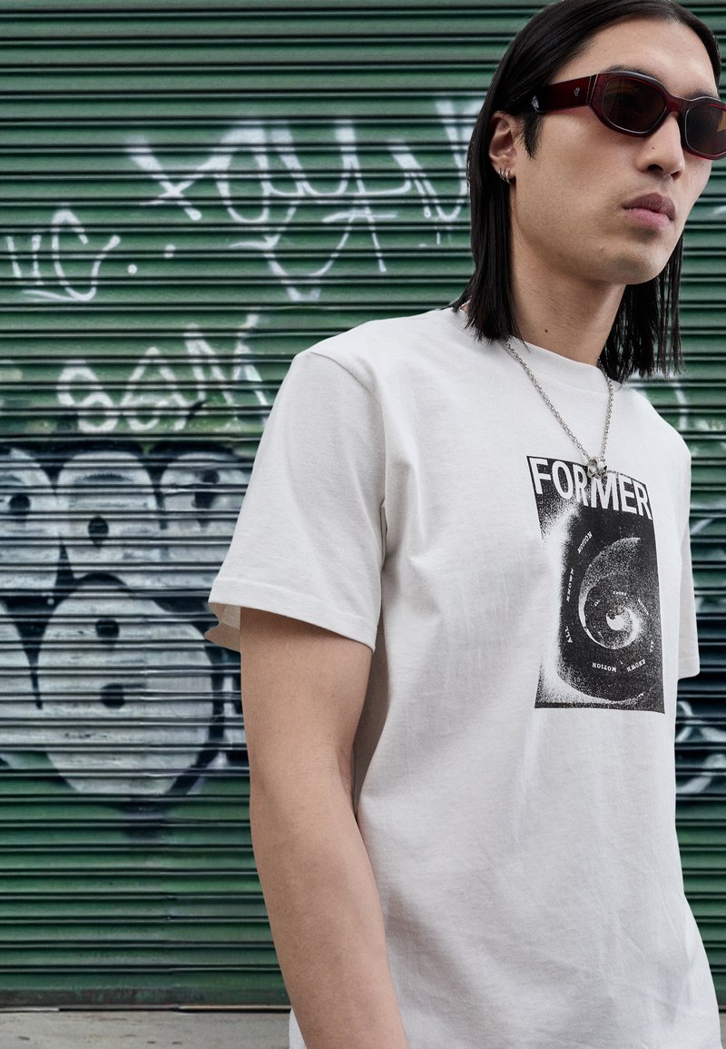 Vit bomullst-shirt med en svart spiralgrafik och ordet "FORMER." Bars med mörka solglasögon och ett silverkedjehalsband.