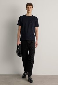 T-shirt noir avec un petit logo sur la poitrine, associé à un pantalon noir et des chaussures noires. Le modèle tient un sac noir.