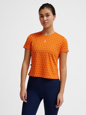 Junge Frau mit dunklen Haaren, die ein oranges Kurzarmshirt mit kleinen schwarzen Fahrradmustern und dunkelblaue Leggings trägt, steht vor weißem Hintergrund.