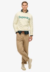 Sudadera beige con el logo verde de "Superdry", llevada sobre una camisa de cuadros verde oscura. Pantalones relajados de color beige claro y zapatillas beige claras.