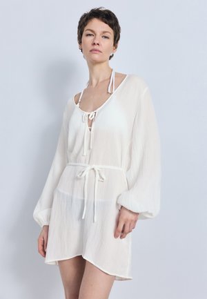 Mujer con cabello corto que lleva una túnica blanca de manga larga con cinturón sobre un top halter blanco, de pie contra un fondo liso y claro.