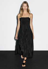 Sort maxi kjole med spaghetti stropper, der har en svævende silhuet og en glat satin tekstur. Detaljerne inkluderer lodrette sømme og en subtil glans.