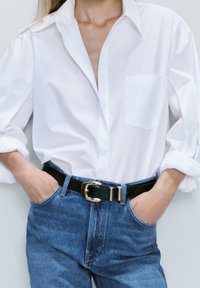 Camicia bianca con maniche arrotolate, abbinata a jeans in denim blu a vita alta, fissati con una cintura nera con fibbia dorata.