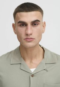Camisa de manga corta, de color verde claro, con cuello, que presenta un detalle de botones en la parte frontal y una tela texturizada. Diseño simple y relajado.