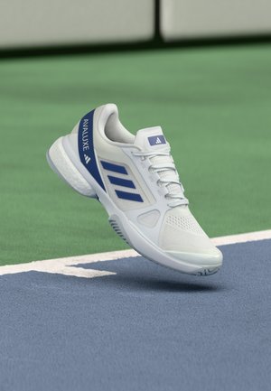 Fehér és kék Adidas Avaluxe teniszcipő lebeg egy kék-zöld pályafelület felett, közel egy fehér vonalhoz.