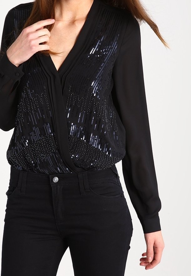 Femme portant un chemisier noir à manches longues orné de sequins foncés et un pantalon noir ajusté, touchant le col avec une main.