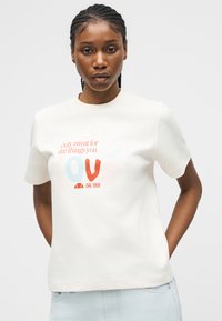 Γυναίκα με πλεξουδάκια στα μαλλιά, που φοράει λευκό T-shirt με κόκκινο και μπλε κείμενο "Μόνο ιδρώτας για τα πράγματα που αγαπάς" και ανοιχτόχρωμο τζιν παντελόνι.