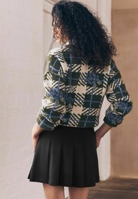 Femme aux cheveux bouclés portant un cardigan à motifs vert, crème et bleu et une jupe plissée noire, se tenant de profil à l'intérieur.