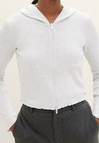 Jasnoszara prążkowana bluza z kapturem zapinana na zamek, o krótkim kroju i przednim zamkiem błyskawicznym; posiada kaptur i długie rękawy. W zestawie z ciemnoszarymi spodniami.